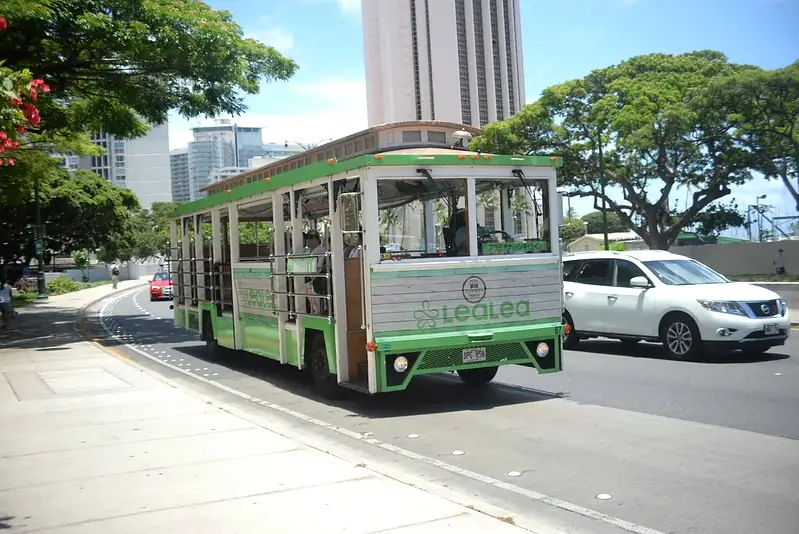 Open-air trolley bus driving on a street in Honolulu with passengers onboar