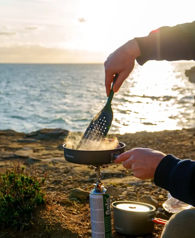 Person using a portable camping stove for outdoor cooking near the ocean